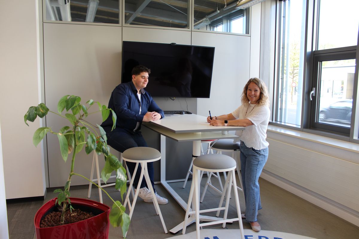 Stehtisch mit mehreren Stehhockern, auf einem sitzt ein Mann. Eine Frau steht am Tisch, dahinter befindet sich ein Bildschirm. Im Vordergrund steht eine Grünpflanze mit rotem Übertopf,