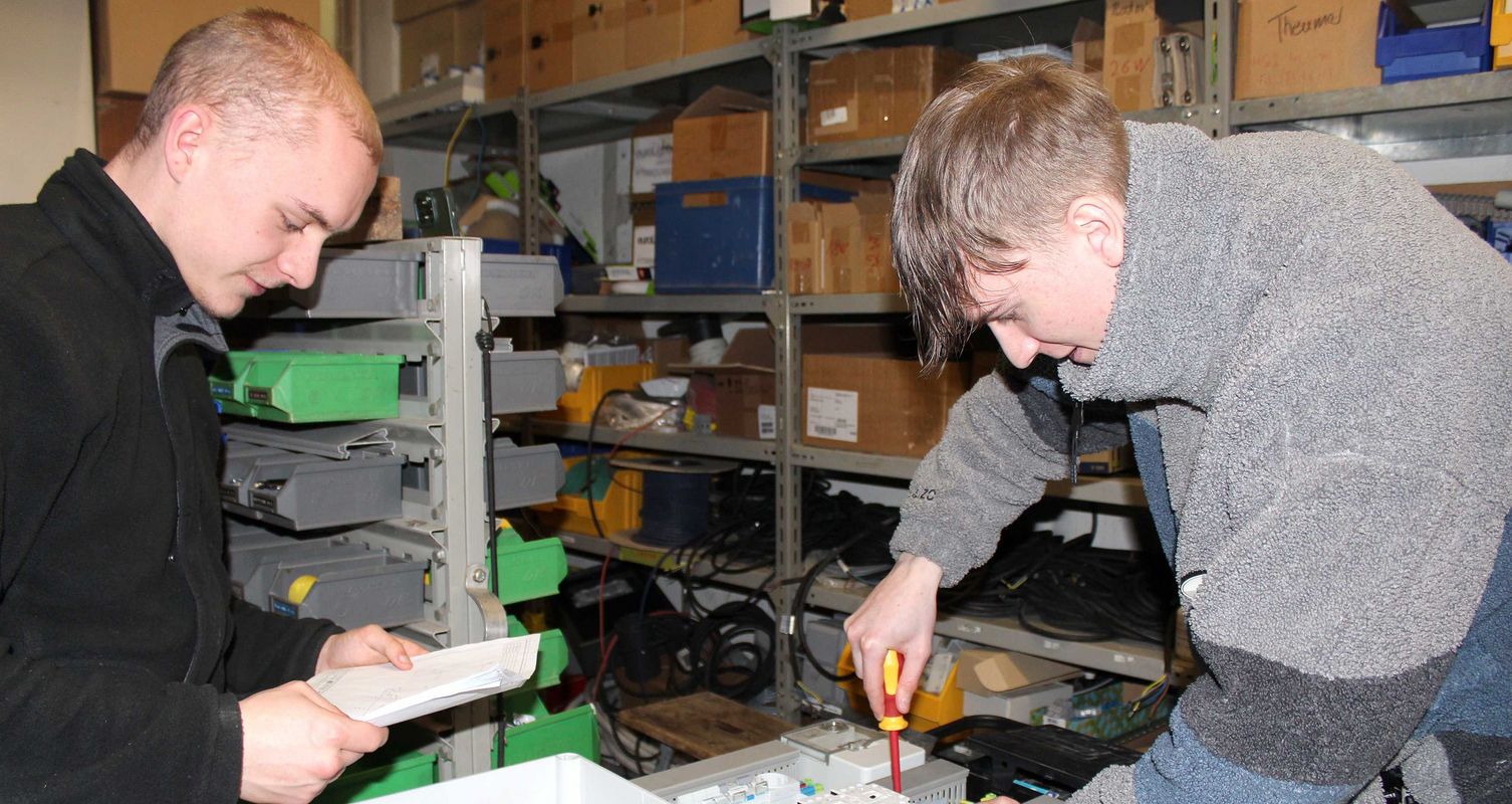 Zwei junge Männer in Arbeitskleidung, einer hält einen Schraubenzieher, der andere liest von einem Blatt. Im Hintergrund sind Regale mit Kartons und Kabeln zu sehen